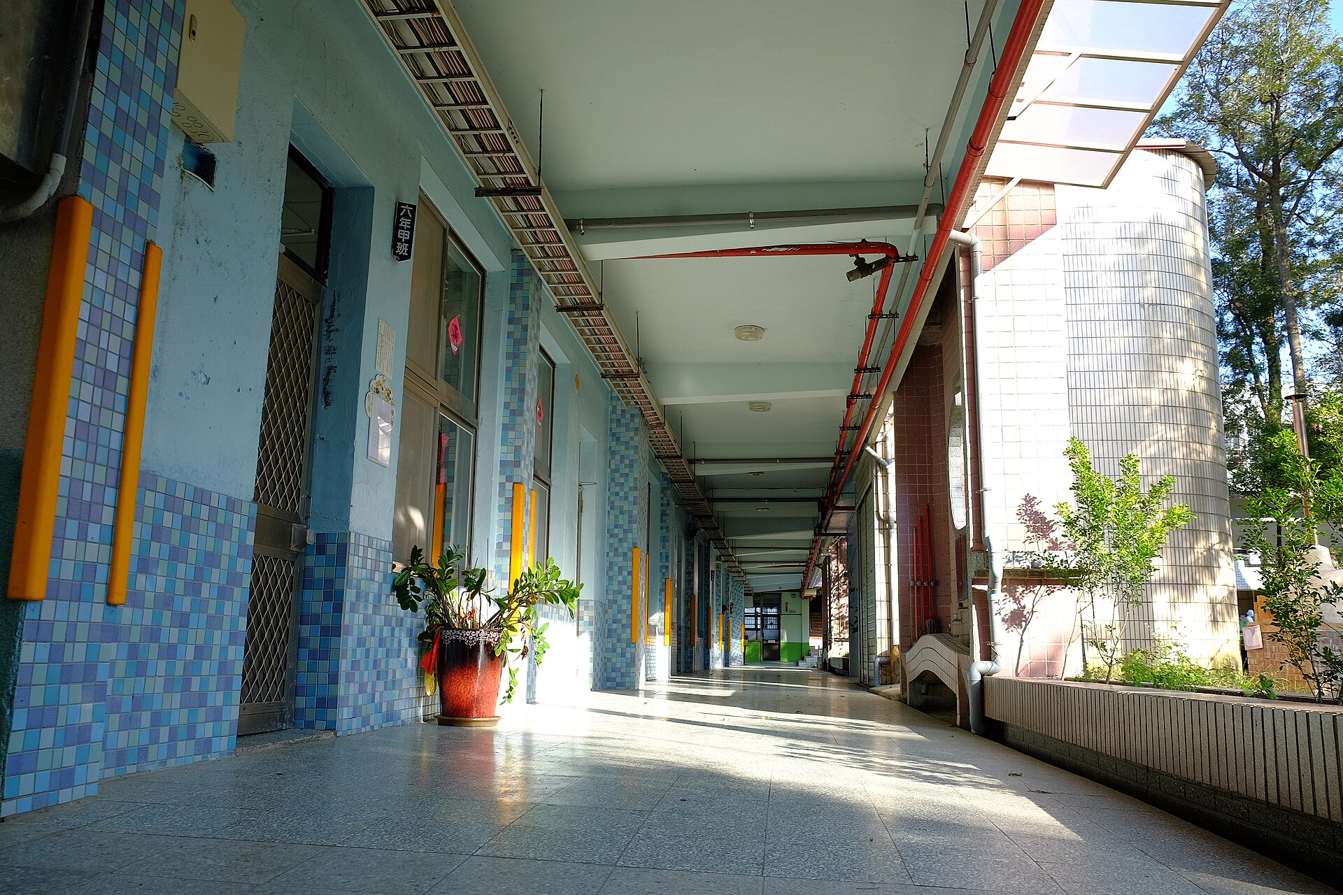 A long, sunlit corridor outside primary-school classrooms, with tiled walls and a row of doors and windows stretching into the distance; no one is in it.