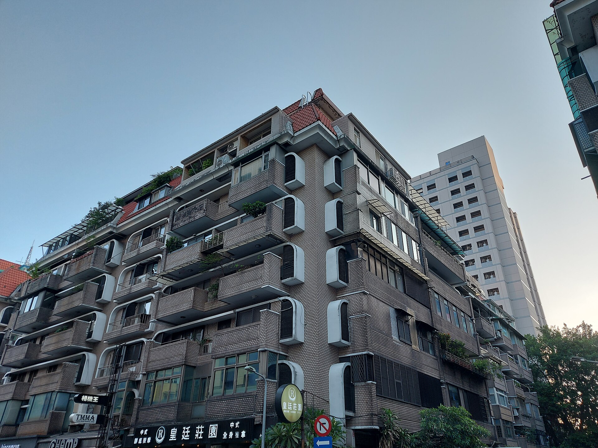 Looking up from a street corner at an old brick-coloured Taipei apartment block with a red-tiled roof and air-conditioners on every balcony, a white tower behind it.