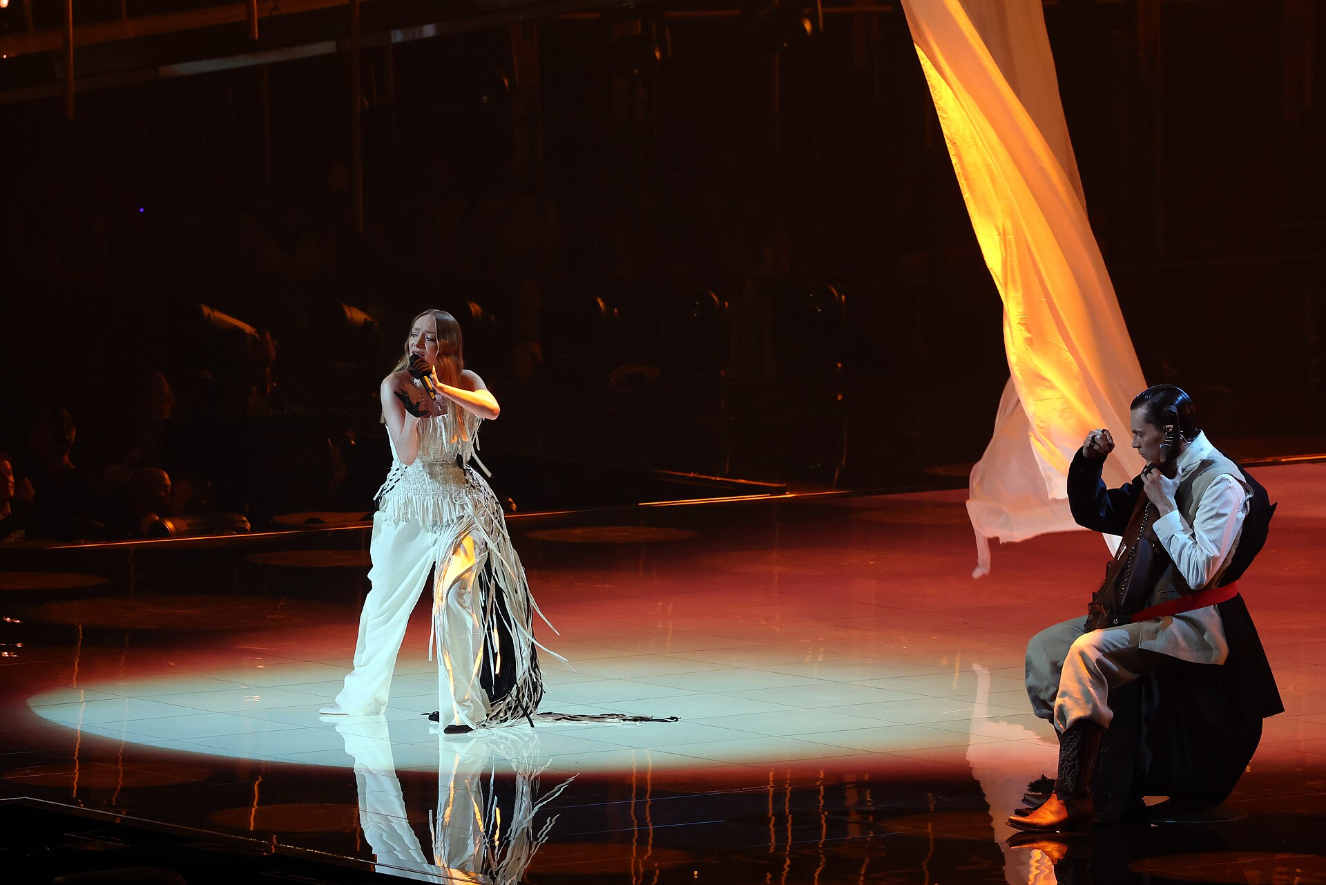 A dark stage: a woman singer in a white fringed outfit stands in a circle of spotlight singing into a microphone, while a man in traditional dress sits playing an instrument on the right, beneath a drape lit orange.