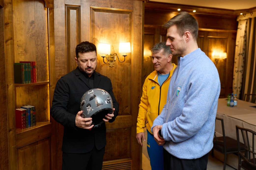 In a wood-panelled room, Zelensky, dressed in black, looks down at a grey helmet covered with portraits; beside him stand a man in a yellow sports jacket and a young athlete in a light-blue sweater.