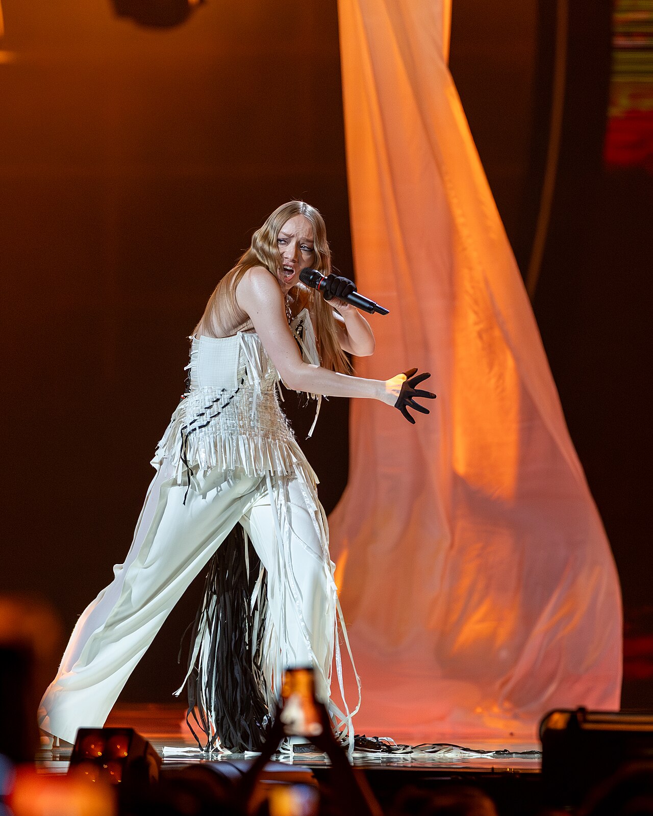A singer in a white fringed outfit and black gloves strides forward singing into a microphone, in front of a drape lit orange, with audience members holding up phones in the foreground.